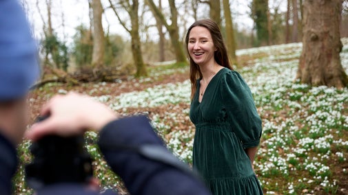 A woman in a velvet green dress smiles and poses for a photo in front of a swathe of snowdrops in the woodland at Stowe.